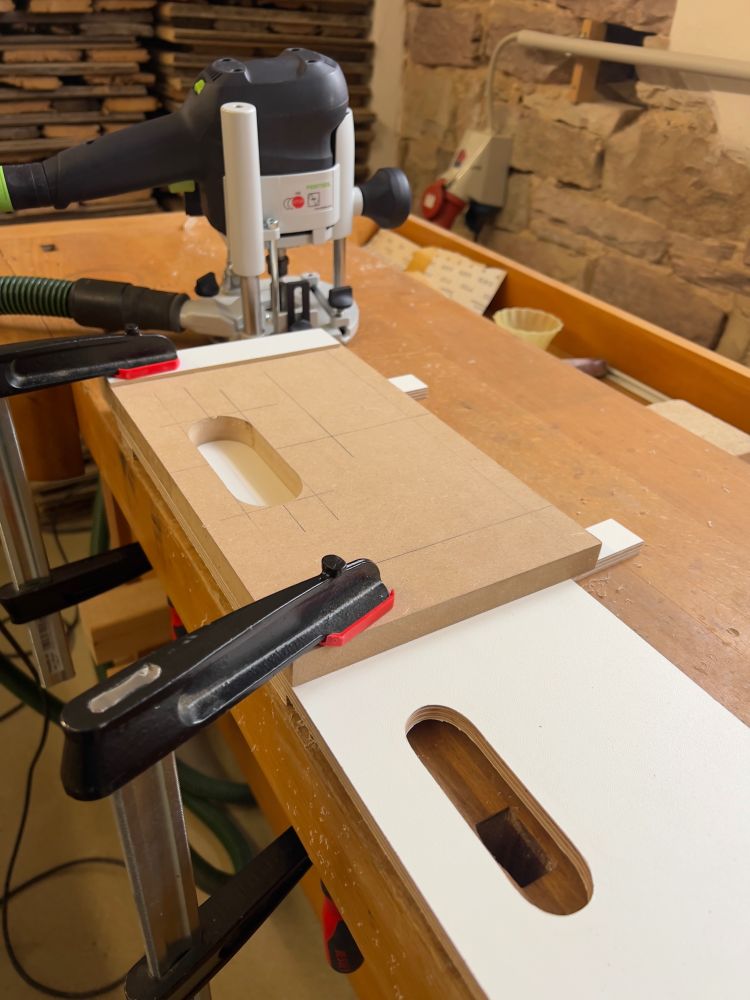 Frässchablone aus MDF mit Schraubzwingen auf eine weiss beschichtete Multiplexplatte aufgespannt. Auf der Hobelbank steht auch eine Oberfräse.