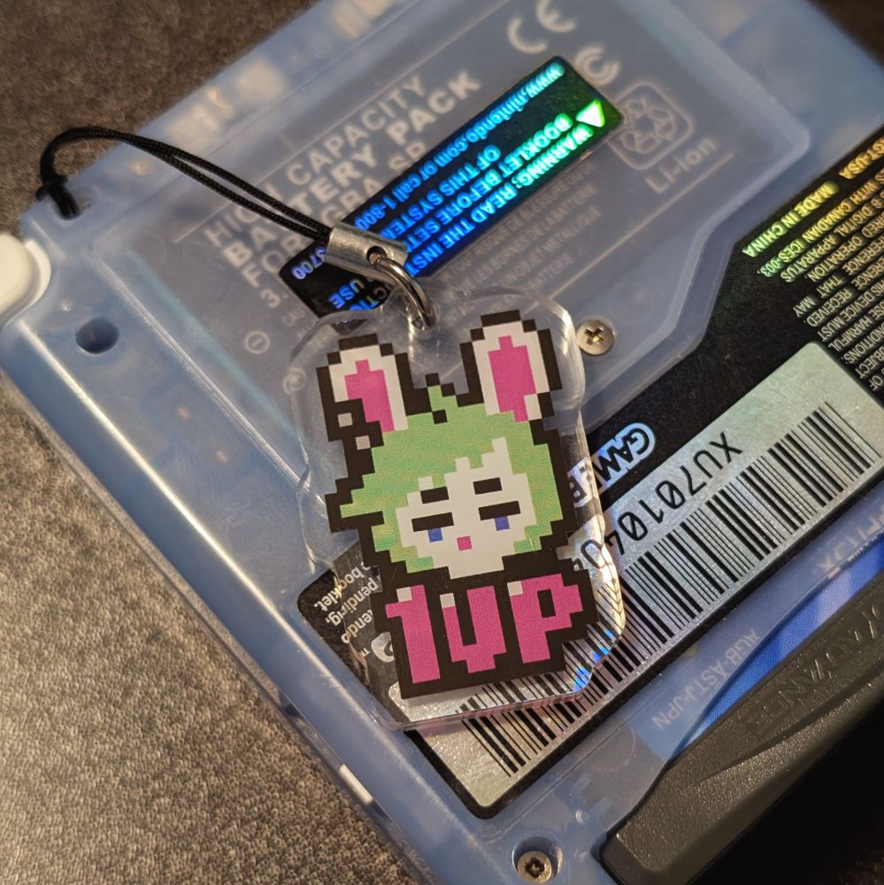 An acrylic keychain of a pixelated rabbit girl head with the phrase "1UP" below her where it's connected to a handheld video game console.