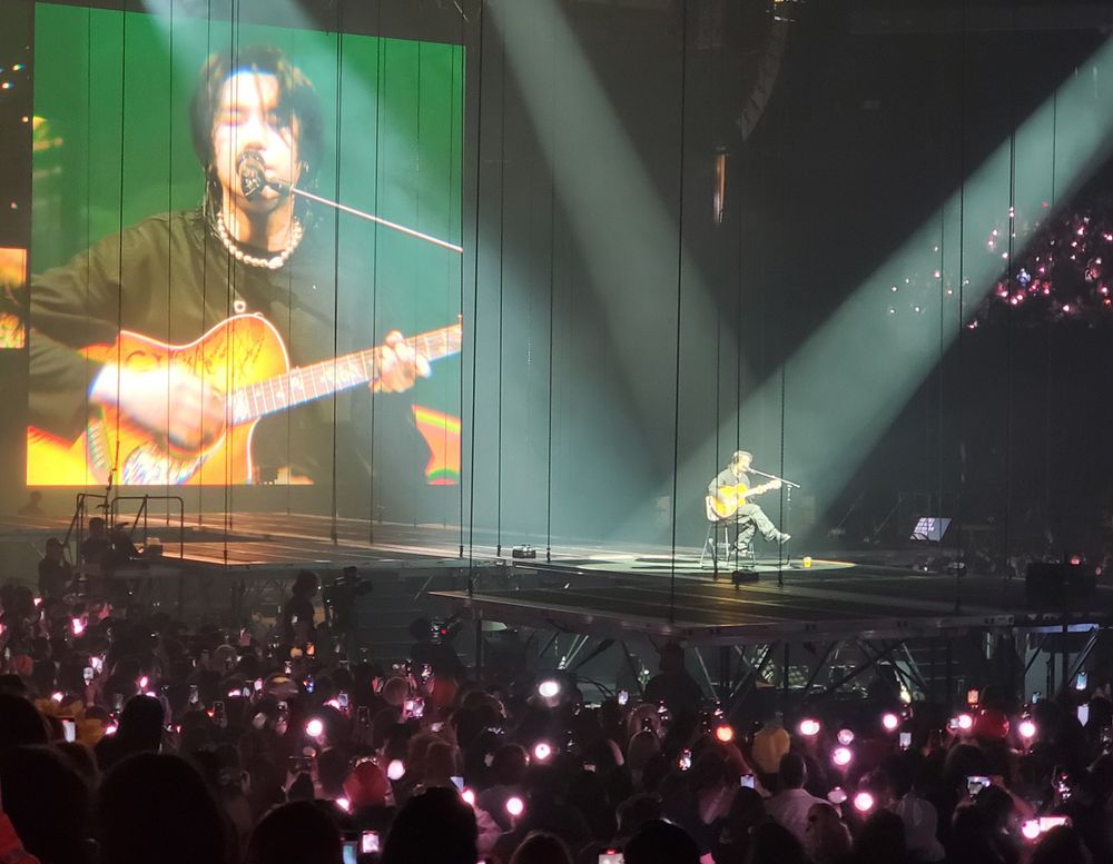 Yoongi on stage for D-Day in Oakland 2023. He's sitting down playing the guitar, and two spotlights catch him with a larger image projected behind him.
