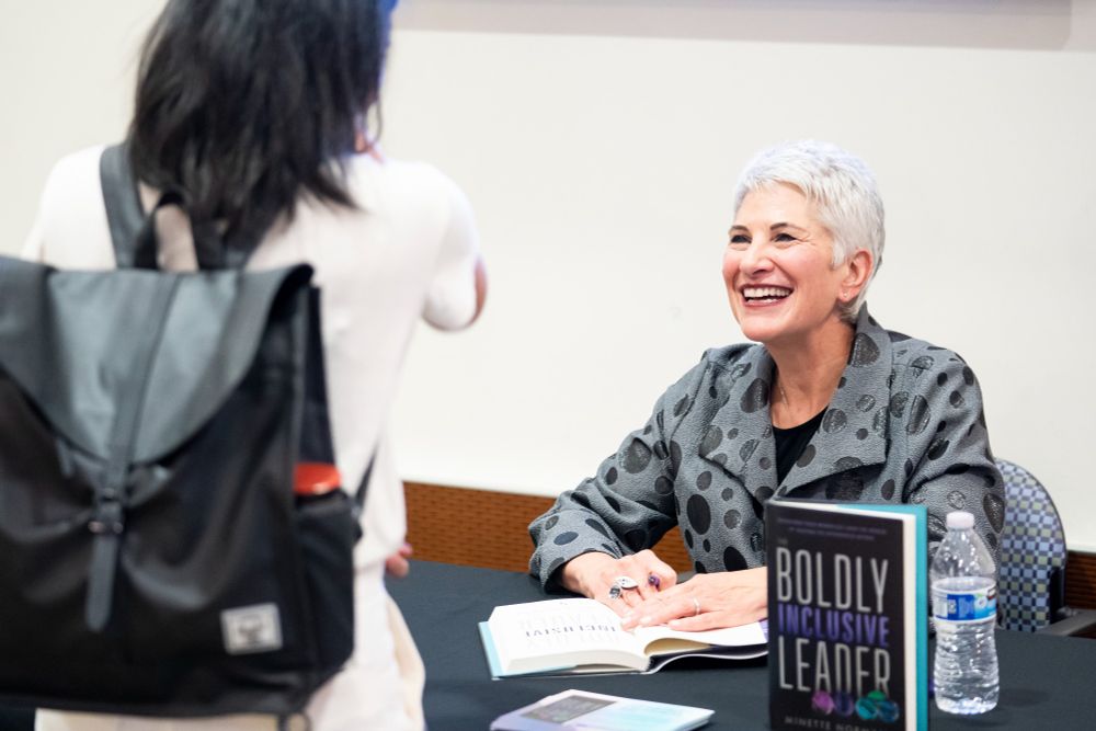 The back of a woman with dark hair, wearing a backpack. She's in conversation with Minette Norman, a white woman with short gray hair wearing a gray and black polka dot blazer. Minette is signing a copy of her book, The Boldly Inclusive Leader.