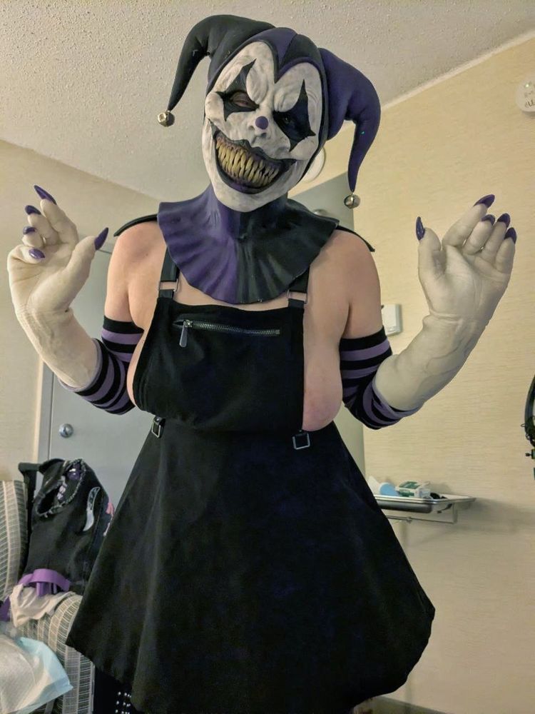 Purple and black femme jester, wearing a black overall dress, smiling wide. Skeeve is owned by @GOOLGRL !