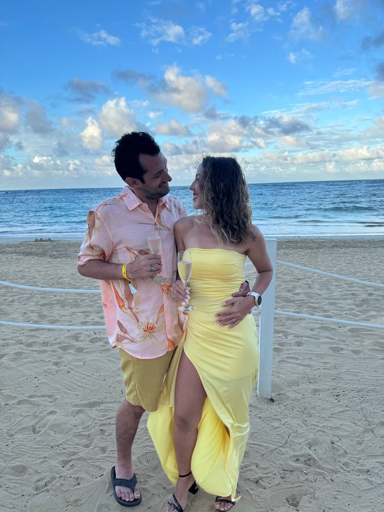 Homem de camisa, bermuda e uma taça na mão abraçado a uma mulher de vestido longo e justo de cor amarela com uma taça na mão 