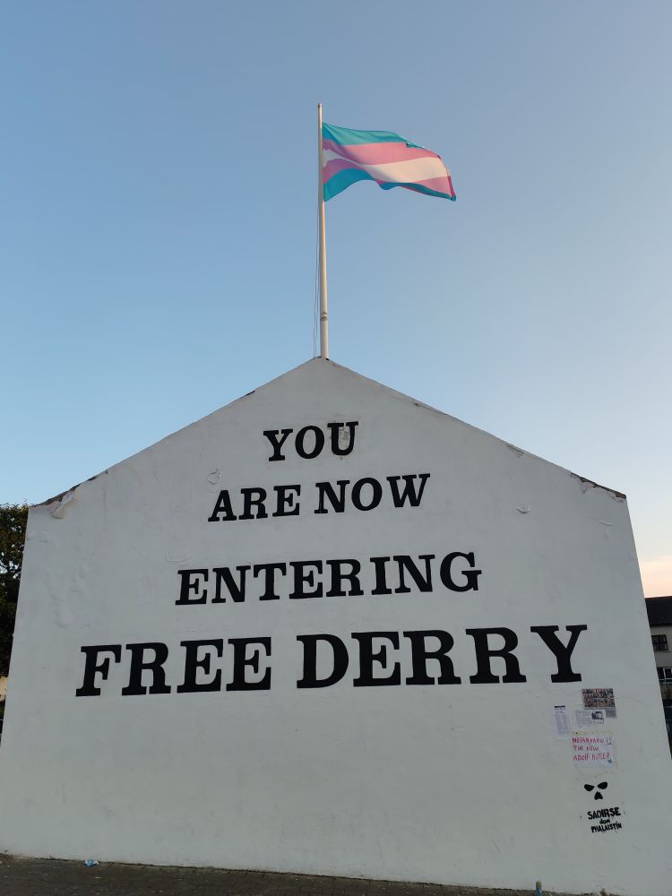 The iconic "You Are New Entering Free Derry" mural in Derry, with the Trans Pride flag flying in the breeze on the flagpole above it. 