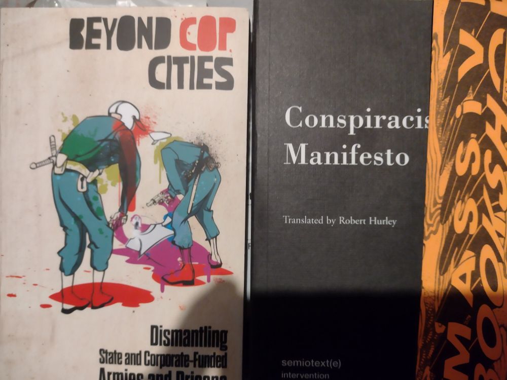 2 book covers (Beyond Cop Cities and Conspiracist Manifesto) and an orange book mark that says Massive Bookshop.

On the cover of Beyond Cop Cities, 2 police officers covered in green paint and blood stand over a man with their guns drawn.

The cover of Conspiracist Manifesto is plain black with the title in white.