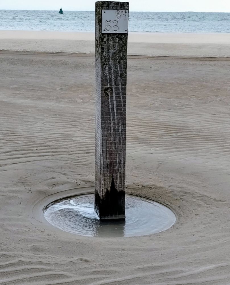A pole on the beach at low tide, a small pool of residual water remains around the pole