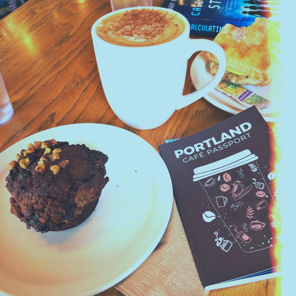 a wooden café table with a chocolate muffin on a white plate, a latte in a white mug, a breakfast sandwich on another white plate, and a copy of the Portland Café Passport