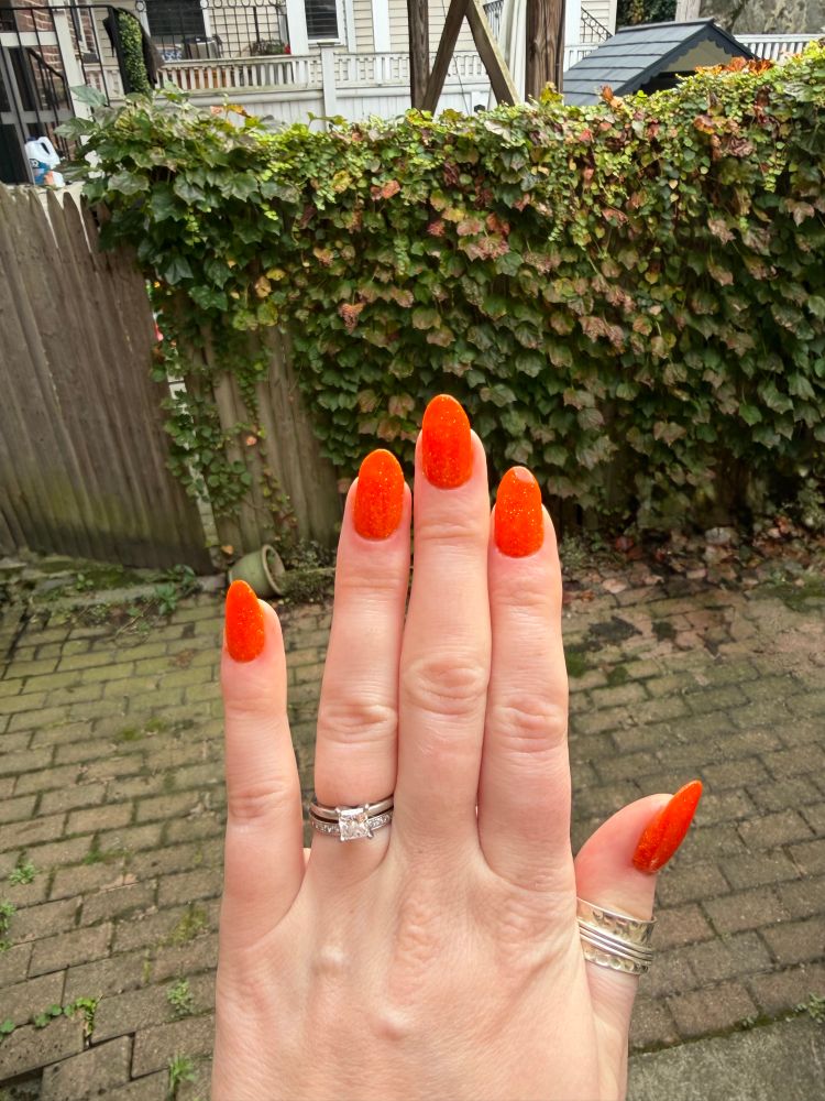 A left hand with orange sparkly almond shaped nails 