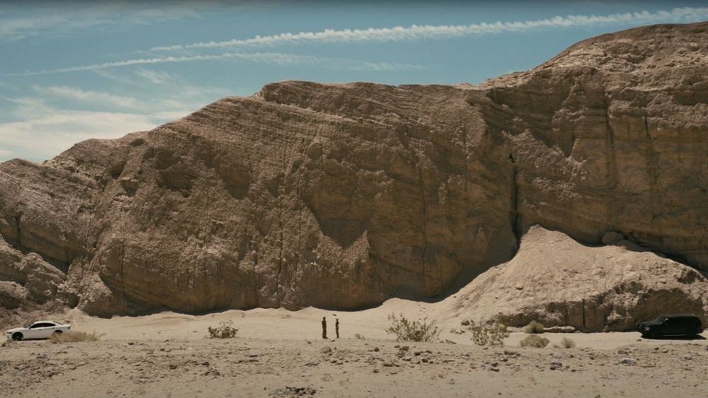Fotograma de la película Una batalla tras otra (2025). Muestra un desierto rodeado de mesetas, con dos figuras de pie en el centro, y un coche blanco a la izquierda.