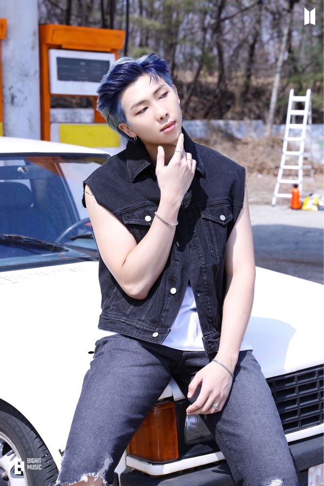 A blue-haired Kim Namjoon of BTS wearing a sleeveless denim vest sitting on the hood of a white car. Photo from behind the scenes of Butter album jacket photoshoot.