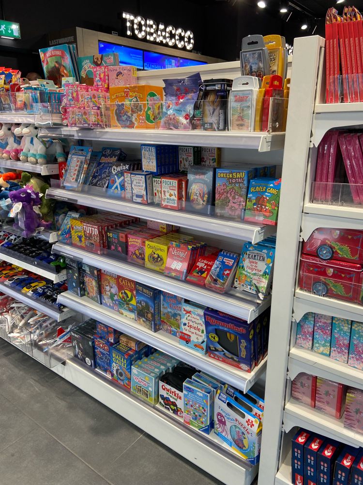 A white shelf of board games next to a shelf of souvenirs in the Zürich airport. A sign reads “tobacco in the background.