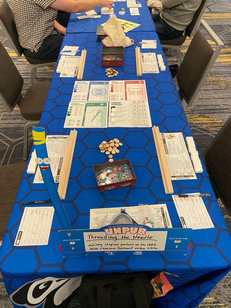 A table with a series of boards and player aids set up on it with mahjong tiles. The table cloth is blue hexagons and the Unpub ready for players sign is raised.