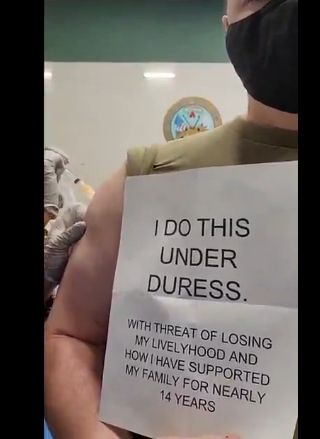 Military man receiving vaccine injection holds paper sign that says: "I do this under Duress. With threat of losing my livelyhood[sic] and how I have supported my family for nearly 14 years".