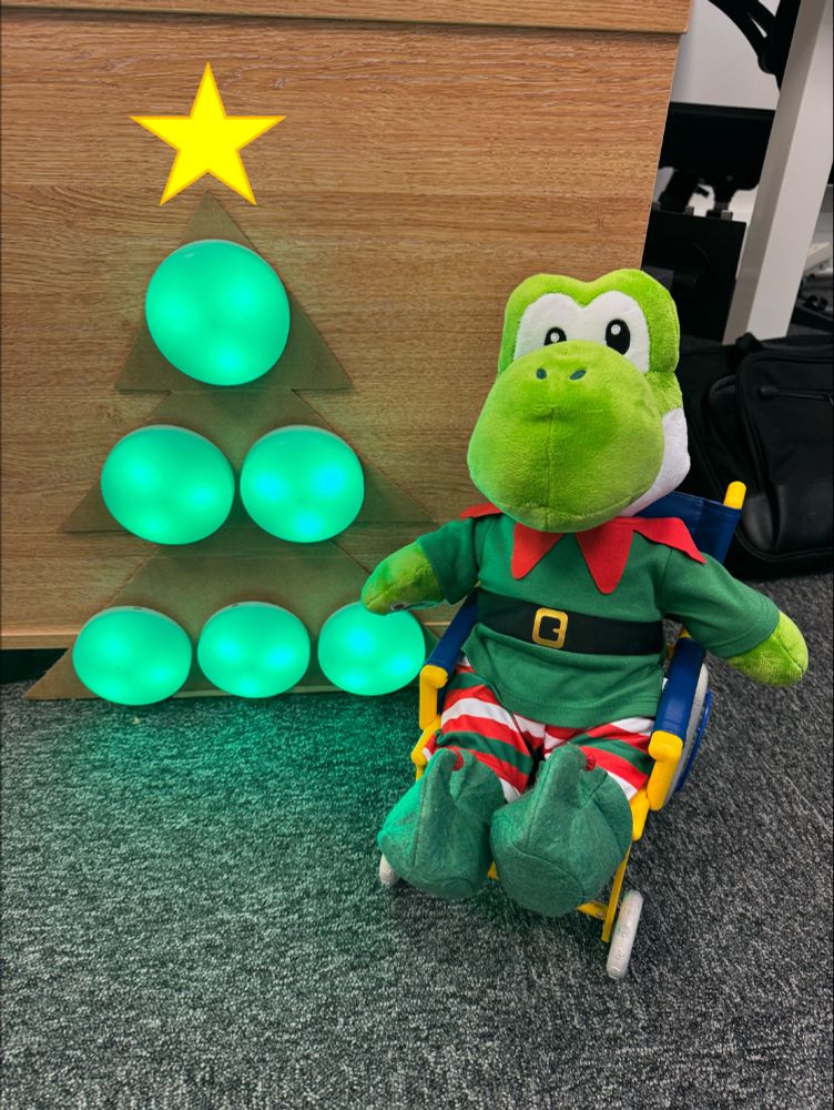 A Yoshi plush sat in a wheelchair, wearing an elf outfit. Next to Yoshi are 6 green Cosmo switches in the shape of a Christmas tree with a yellow star above them.