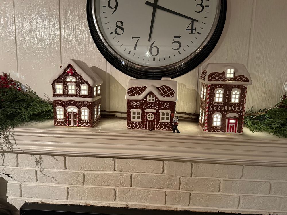 Three “gingerbread” holiday display houses. The two on ends have lights on. The middle house does not. A figure stands in front of the corner of the house…