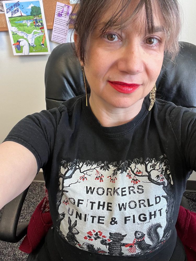 Brunette Woman with bangs sitting at desk. Behind her is a picture that says eat the Rich. Her black t shirt has forest creatures on it with the words “workers of the world unite and fight”