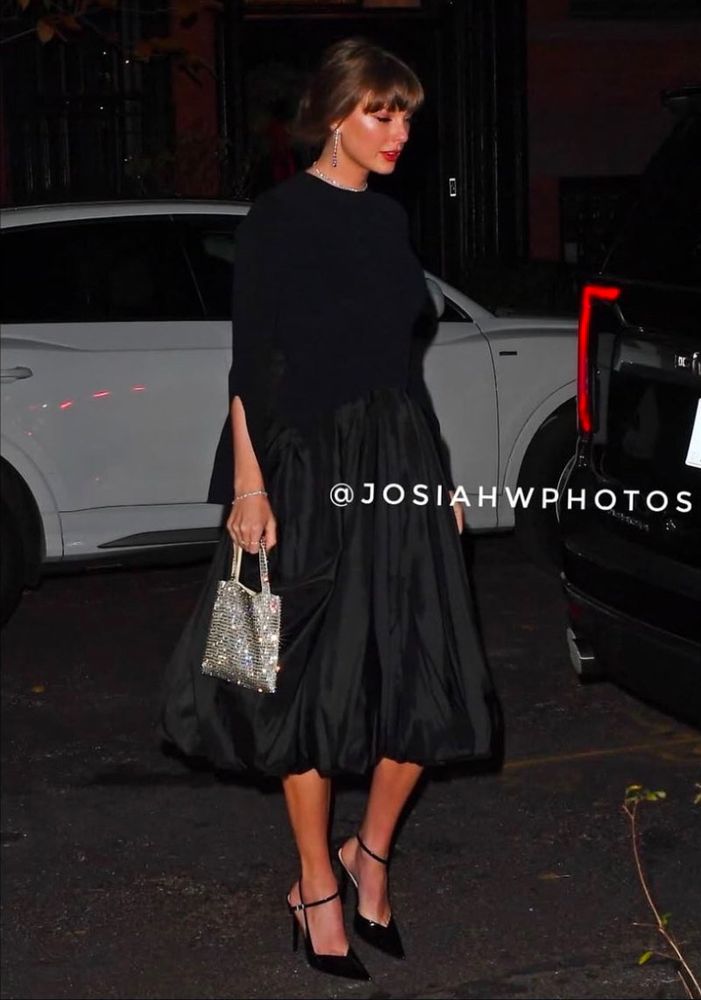 taylor swift wearing a black top, black long skirt, black heels and a sparkly silver bag, with her hair in a bun 