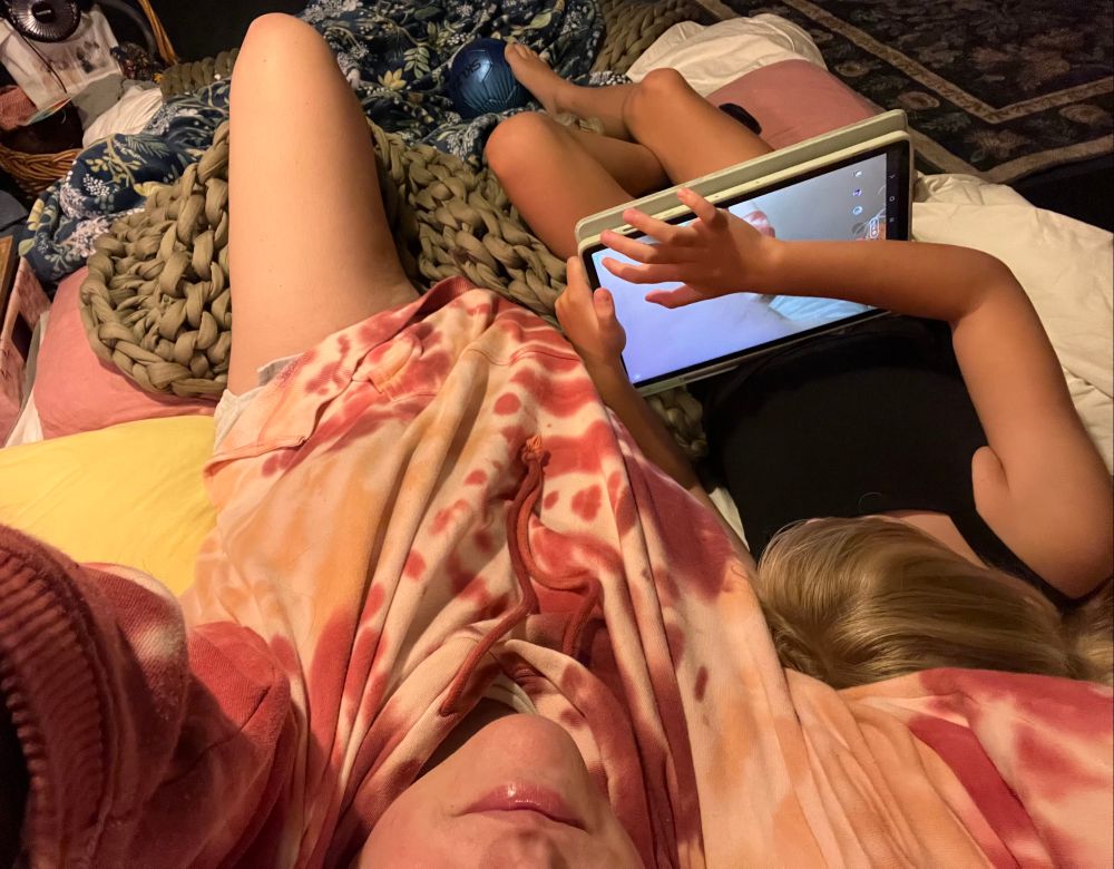 clue overhead selfie laying in her nest next to blonde kid who’s face isn’t visible. kid is on a tablet. there’s a small soccer ball at the kids feet 