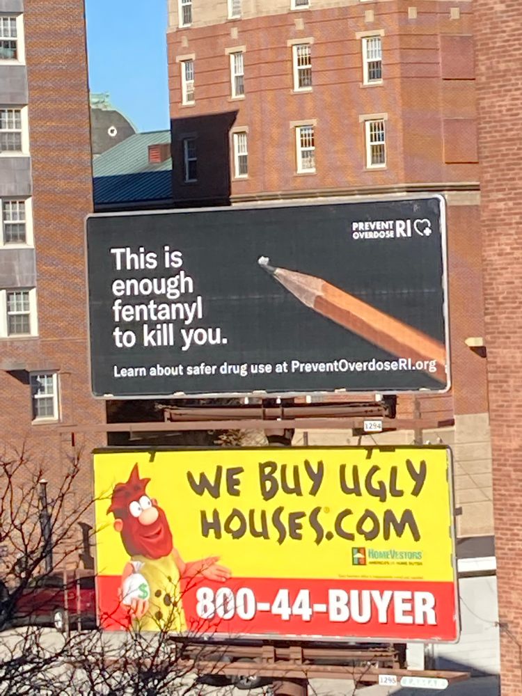 A billboard with a black background a large sharpened pencil with a small white substance on the tip and the white words:
This is enough fentanyl to kill you.
In smaller font:
Learn about safer drug use at PreventOverdoseRI.org.
Another billboard below has a bright yellow background and a cartoon image of a bearded man and in all upper case says: We buy Ugly Houses.com and 800-44-buyer.
Behind the billboards are two brick buildings and a small amount of blue sky.