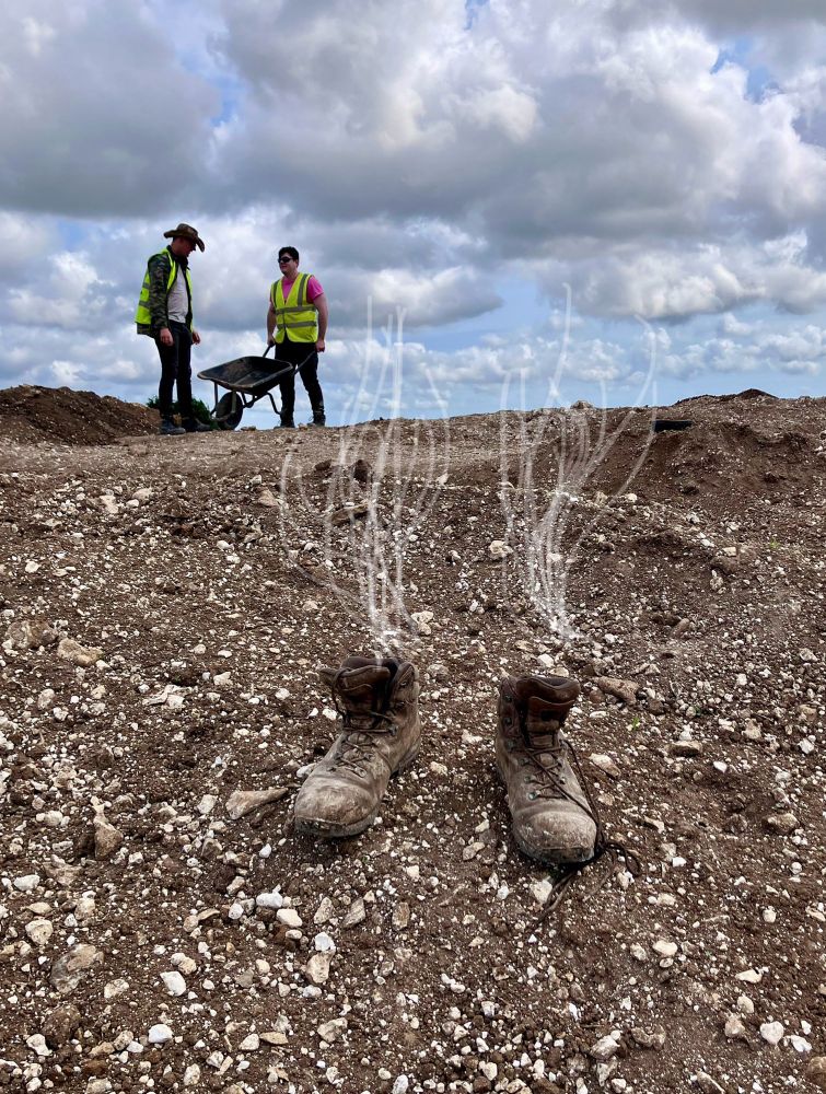 A pair of muddy boots are planted on the spoil heap as if the owner has just evaporated. Two archaeologists are in hi-vis jackets are in the background