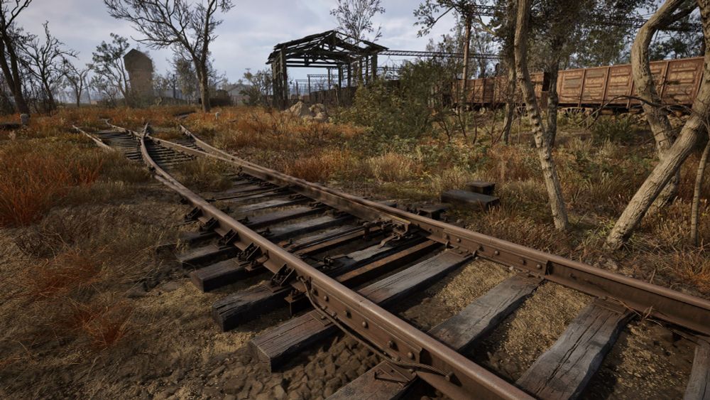 A screenshot from STALKER 2 videogame. A old train track switch with wooden ties is in the foreground. The ground is covered in dense grass. A few train cars and station buildings are seen in the distance.