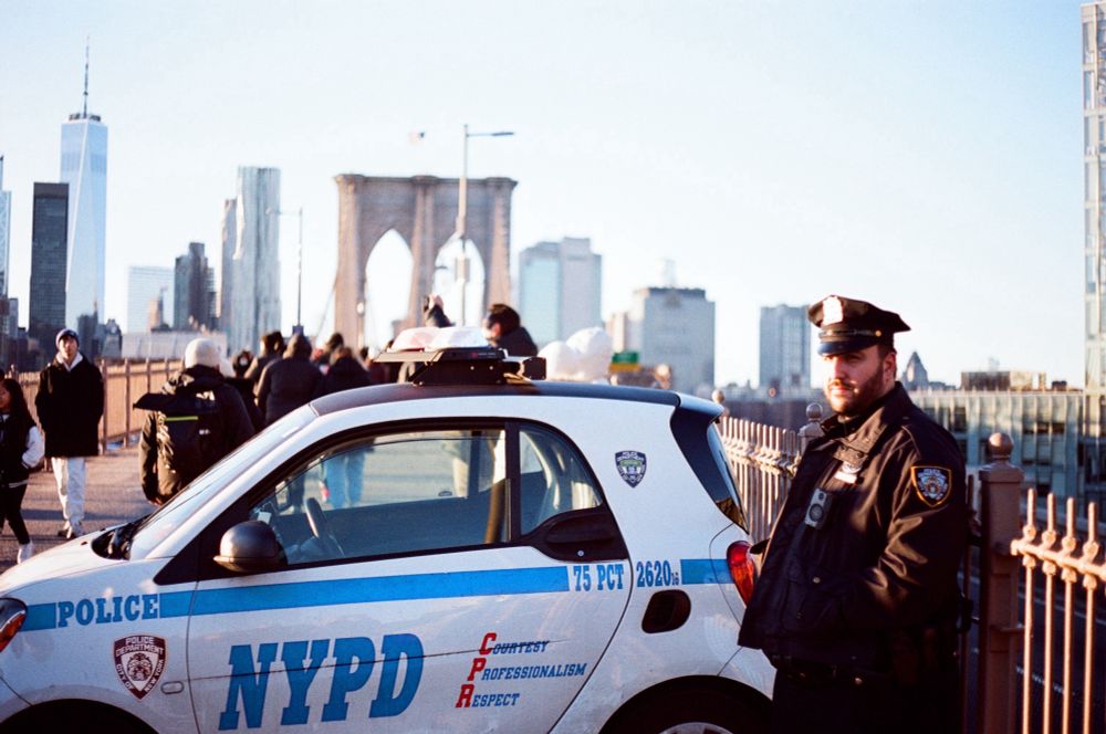A NYPD code stands in front of a small e-cruiser parked on the Brooklyn bridge, squinting towards the camera in the late afternoon sun. The bridge and city, including the freedom tower, visible in the background. ACAB
