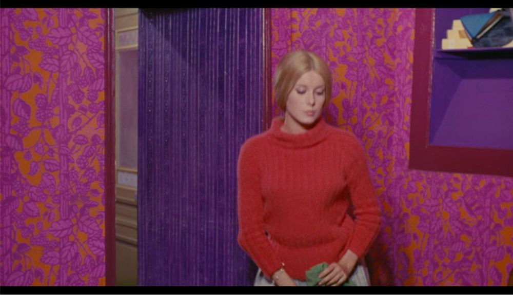 a blonde woman is in the act of sitting down so she's kinda blurry. the walls behind her are orange and pink patterned wallpaper (!) and the door is purple (!!) 
