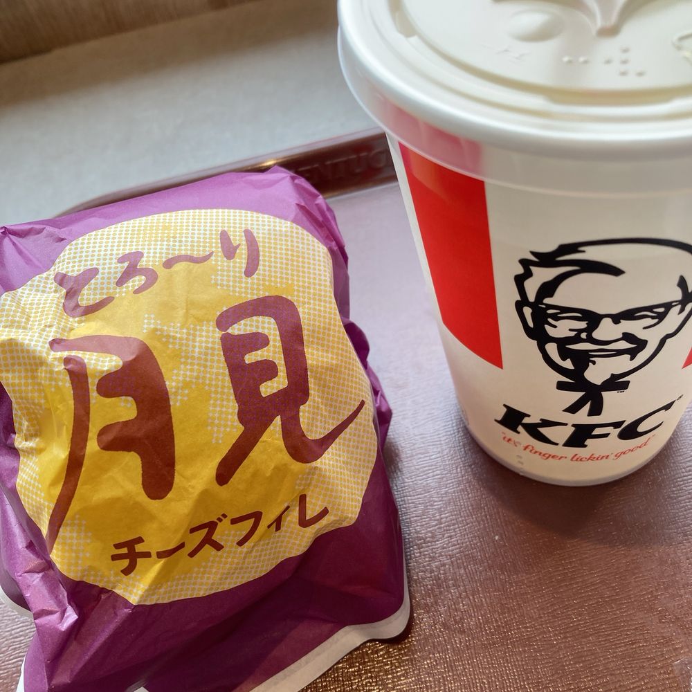 ケンタッキーフライドチキンのとろ〜り月見チーズフィレバーガーの包み(紫地に月をイメージした黄色の円が描かれている)とKFCロゴとカーネルサンダースの顔が描かれた飲み物のカップが並んでいる写真！