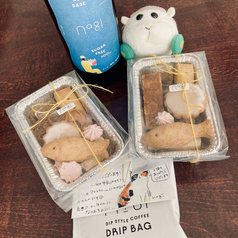 nagi coffee&bakeさんのカフェオレベース瓶(ガラス瓶に青い海を走るボート、それを眺めるバケツいっぱいの魚を抱えた人がデザインされたステッカーが貼ってある)と、通称シャケ弁クッキー詰め合わせ(魚の形のクッキーが一番上に乗っているため)2個、おまけのドリップコーヒーパッケージを並べている。それを見つめるシロモちゃん。