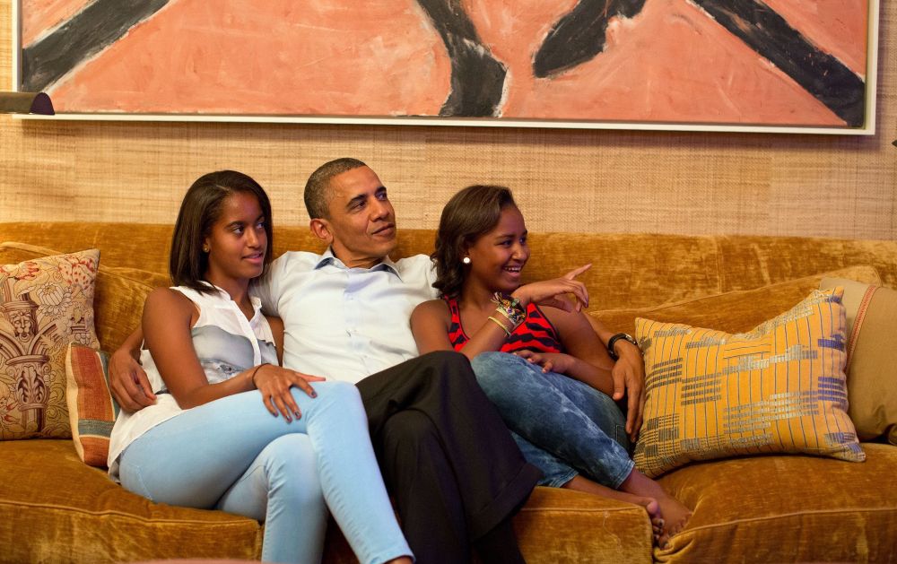 President Barack Obama sitting on a couch with Sasha and Malia Obama, smiling in a room decorated with a large painting and patterned pillows.