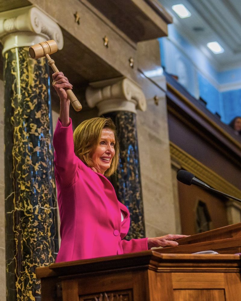 Speaker Nancy Pelosi smiling and waving a gavel while standing at the rostrum of the House of Representatives.