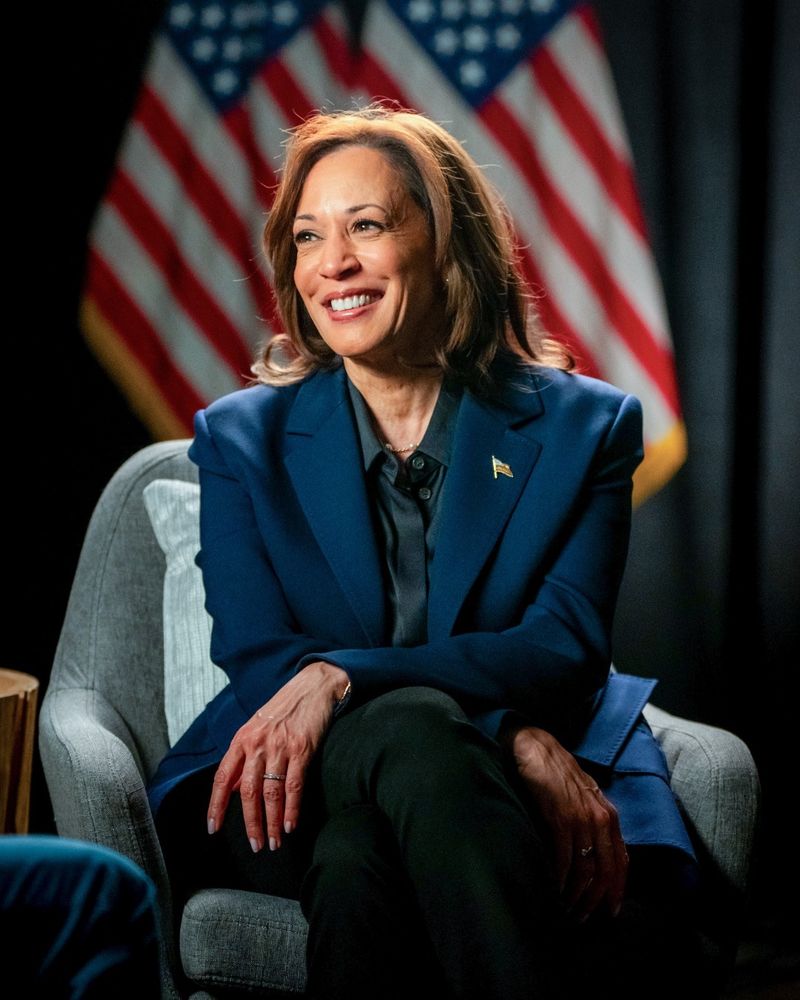 Vice President Kamala Harris sitting with a smile, wearing a blue suit, against a backdrop of American flags.