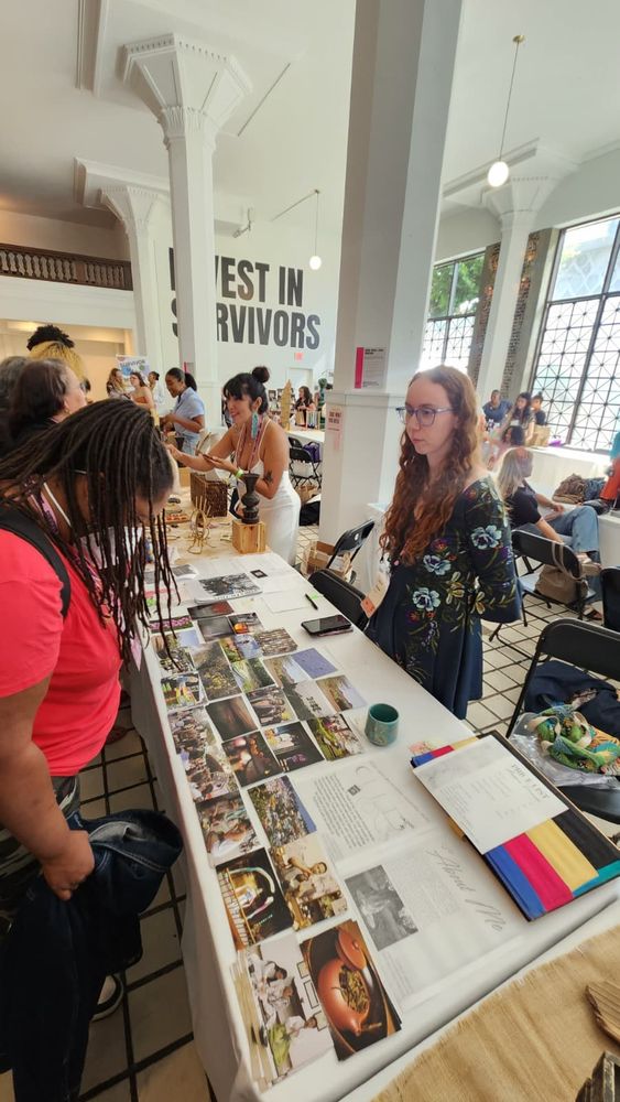 Anesce Dremen, a red-head with curly hair wearing a teal dress and blue glasses, stands behind a table with white cloth where 30 postcards are strewn across the table. A customer wearing a red shirt looks down at the postcards. Additional vendors, all survivors, sell their goods in tables behind the back. Bold letters are painted on the back wall and read: INVEST IN SURVIVORS. 