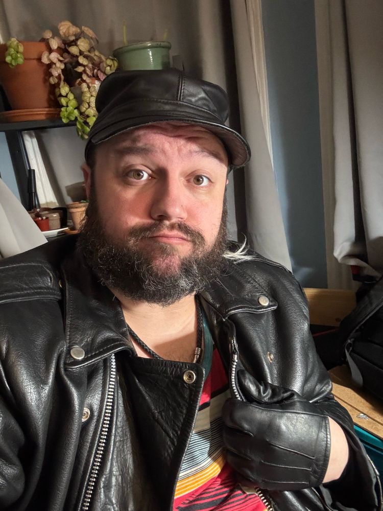 Selfie of Marrok, making a bit of a frown while wearing their heavy leather jacket, gloves, and hat.