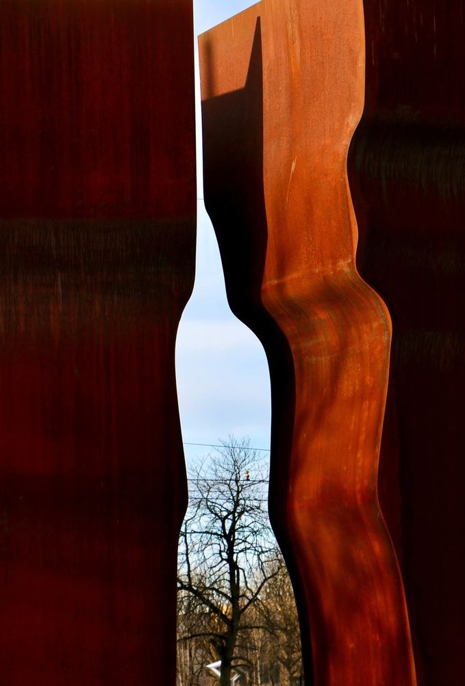 Detail einer korrigierten Stahlskulptur. Zwei leicht gewellte, sich nach oben verbreiternde und mehrere Meter Höhe abstrakte Formen. Die Rechte von der Sonne angestrahlt, wodurch das rostrot zur Geltung kommt, die linke im Schatten. Zwischen den Stelen öffnet sich der Blick auf einen Baum im Hintergrund.