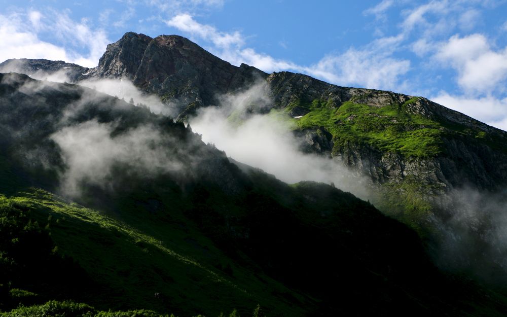 Mehrere Alpenhänge, teilweise von der Sonne beschienen und grün leuchtend, teilweise im Schatten liegend und nahezu schwarz. Darüber im oberen Viertel ein blauer Himmel. Aus den sonnenbeschienenen Hügel steigen Nebelschwaden.