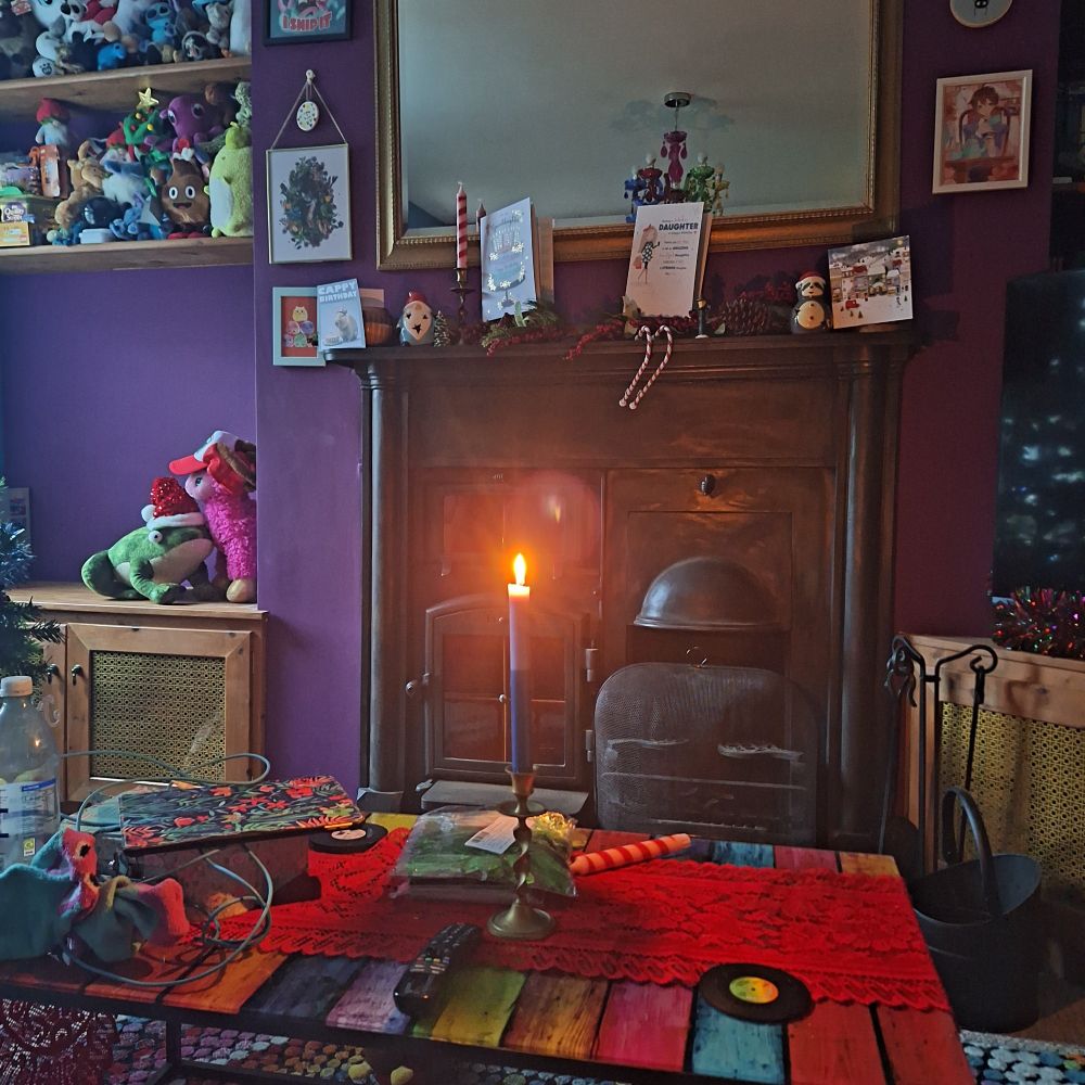 A single candle burns on a cluttered glass table in a darkened room in front of a large metal hearth. Everything is decorated in rainbow colours.