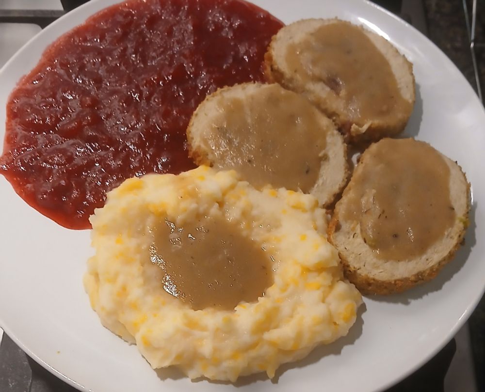 Plate of food featuring slices of vegan roast and cheesy mashed potatoes smothered in gravy, with a large side of homemade cranberry sauce.