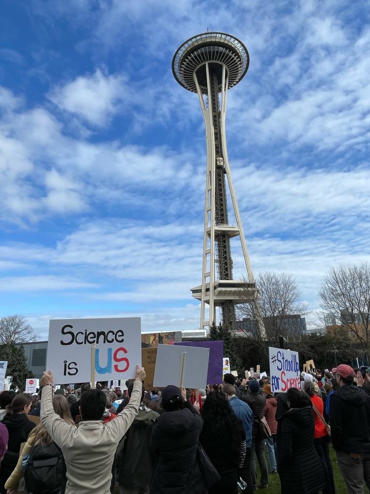 With the Space Needle and a partly cloudy sky as a backdrop, people hold signs at today’s rally. One says “Science is US.” Another reads “#StandUp for Science.”