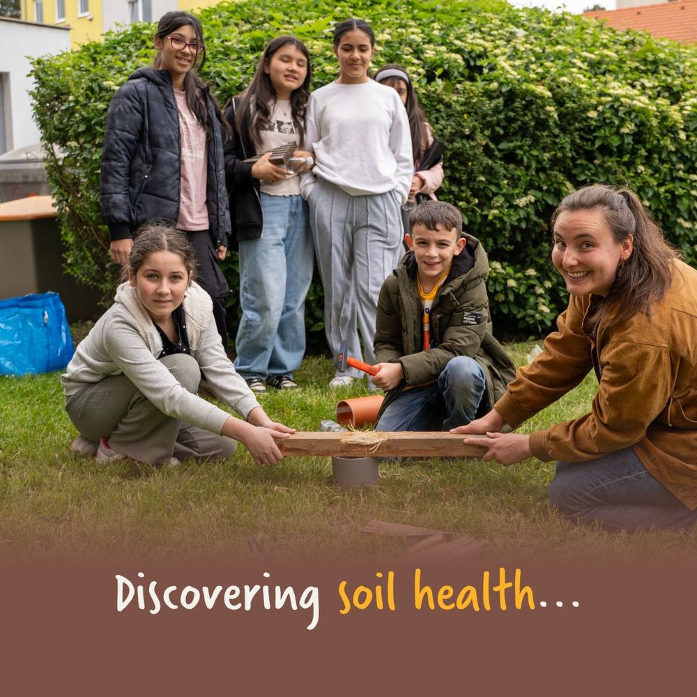 A group of kids and an adult are outdoors, smiling and engaging in a hands-on activity about soil health. They are gathered around a wooden plank and soil tools on the grass, with a big green hedge in the background. The text on the image reads, "Discovering soil health..."