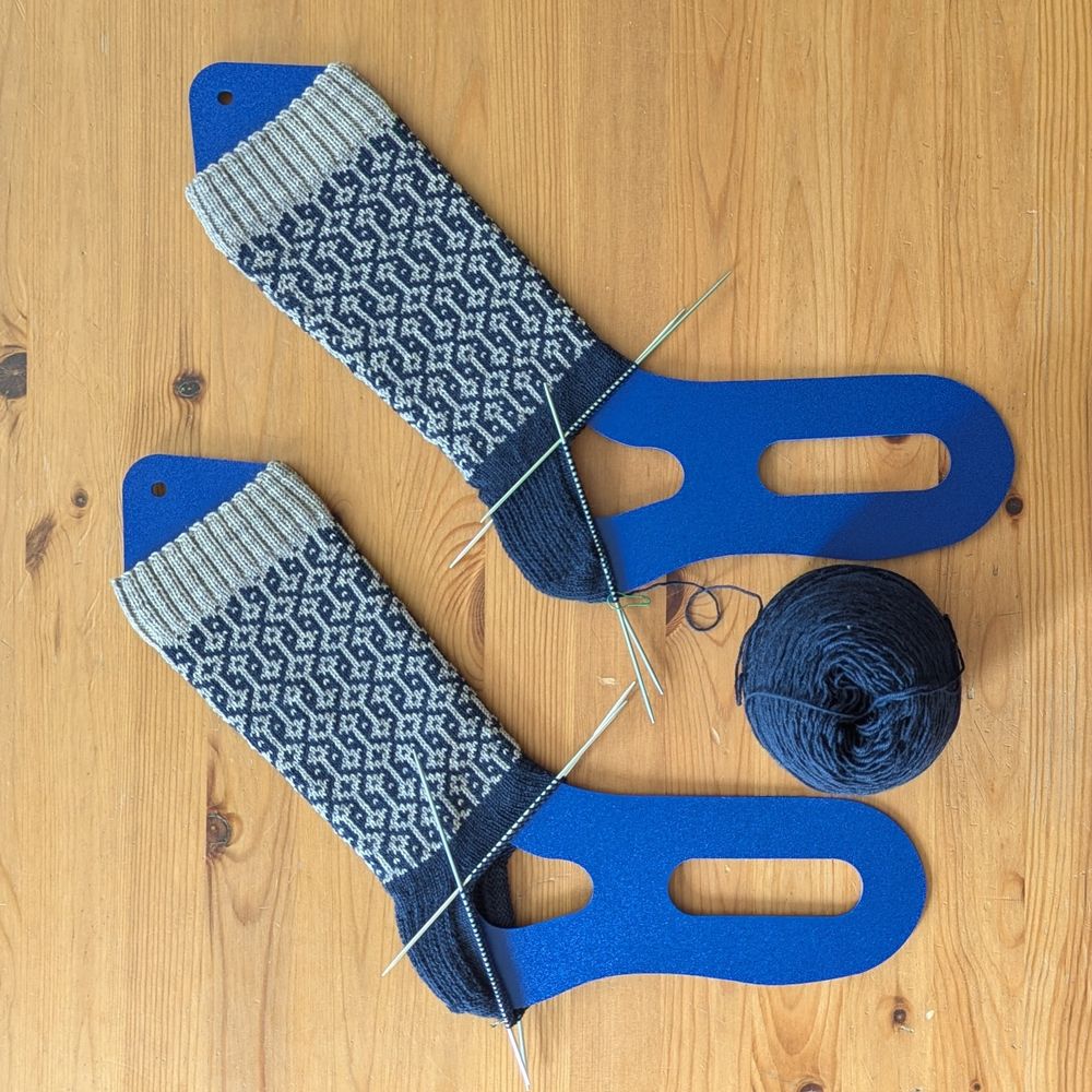 A pair of partially-completed socks with a gray cuff and a navy-and gray colourwork pattern on the leg. The heels are navy. Both socks are on plastic blockers for display and both of them only have the cuff, leg, and heel knit so far.