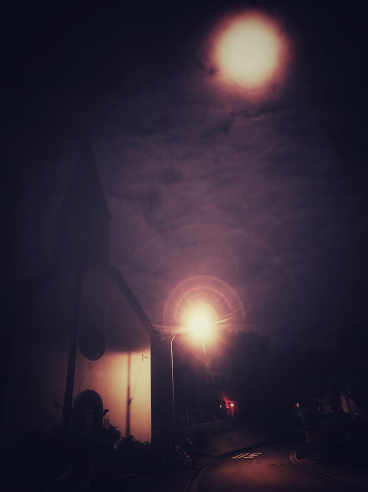 Nighttime by a village church, the moon shines bright behind the clouds.