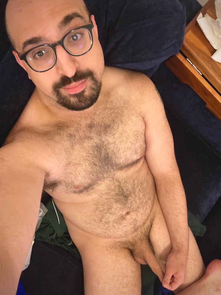 A gay Arab-American man sits on a deep indigo blue couch, completely naked. He is bald, with a dark brown goatee on his chin being the only hair on his head. He wears thick, round, dark blue glasses, and is looking towards the camera smirking. Below his waist, he is tugging his foreskin and stretching it out, about an inch or so past the head of his penis. 