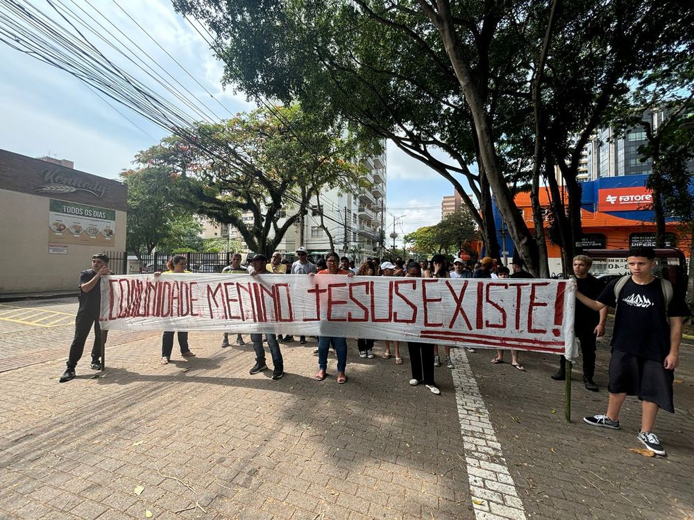 Povo na rua pelo Ato contra o Despejo da Comunidade Menino Jesus, de São José dos Campos (SP). Faixa do bloco, ocupando a avenida, escrito: "Comunidade Menino Jesus Existe!", referência porque a comunidade não é reconhecida pelo povo da cidade devido ao Estado praticar uma política de isolamento da comunidade.