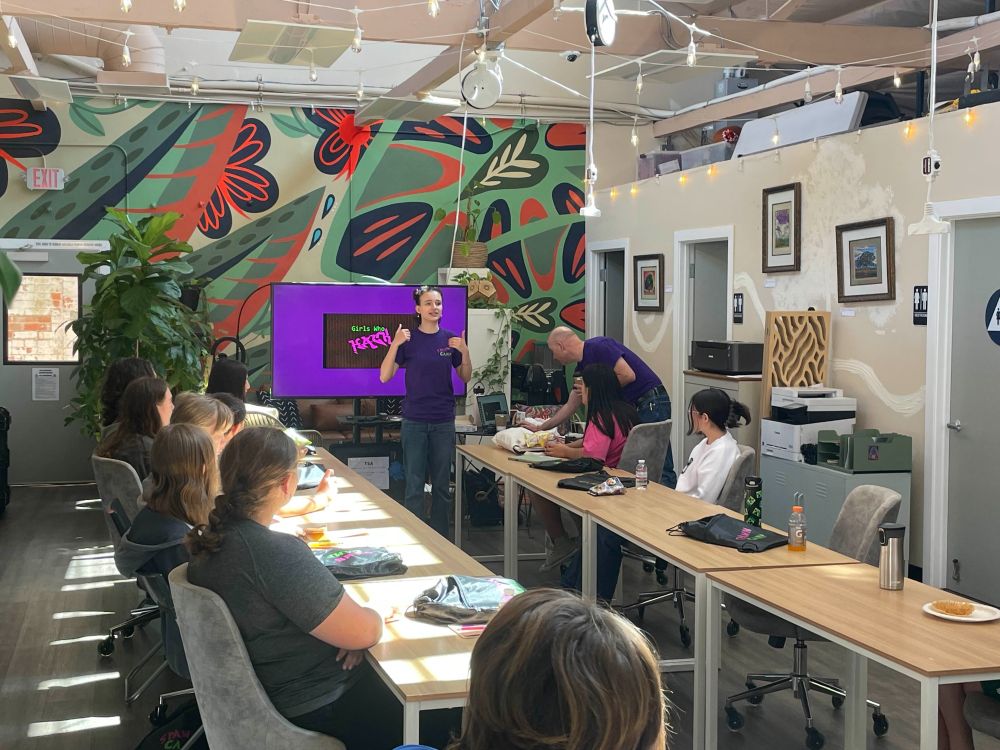 A young woman standing in front of a group of women and a presentation teaching and explaining cybersecurity