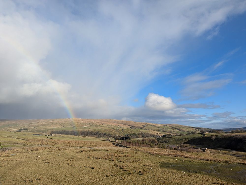 High moorland landscape with blue sky on the right, cloud on the left. A rainbow touches the ground under the cloud 