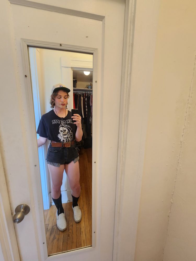 Alex Haus of Decline wearing off white Jays baseball cap, Black t shirt with grim reaper holding severed penis titled Dick Ripper. This is by Rory Blank Bonejail. She is wearing black cut offs, black socks and white vans.