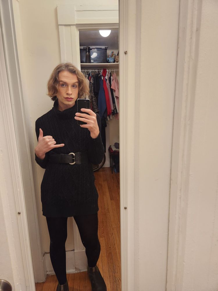 Alex Haus of Decline dressed in a sweater tied down with a waist ceinture and pulled down so as to resemble a dress. She is wearing black stocking and black boots with green trim. Contrary to the posts message she has a nonchalant expression but is doing the "Rad" hand geature with her thumb and pinky extended in opposite directions.