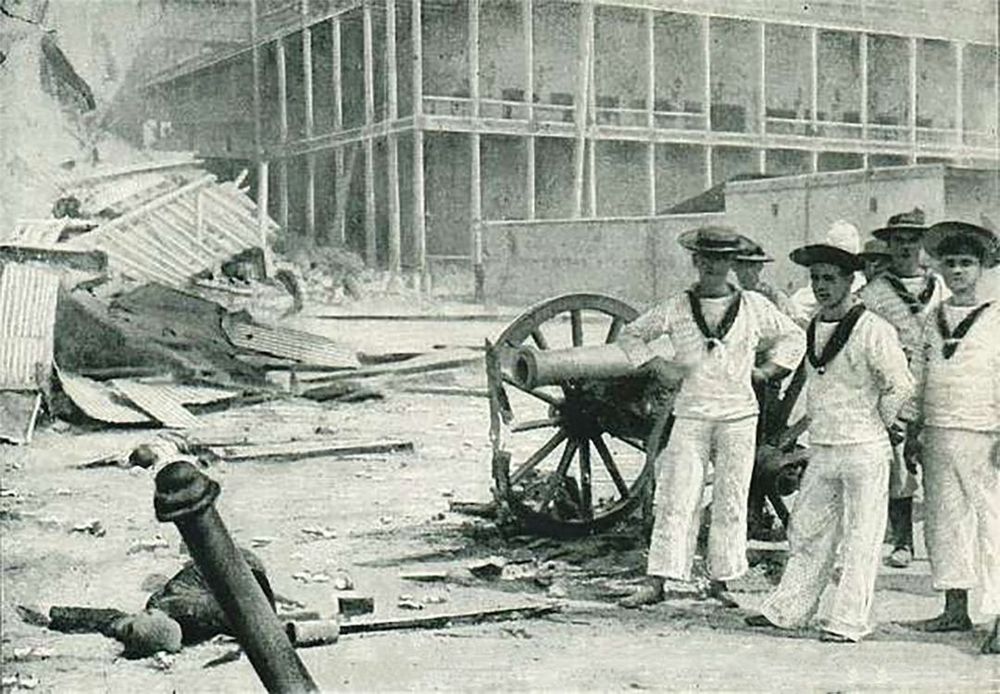A black and white photo showing British sailors in white uniforms standing beside a small field gun amid rubble outside a large wooden building. Fallen cannon barrels, shattered corrugated metal, and debris fill the foreground, evoking the immediate aftermath of the bombardment. Image: Public Domain