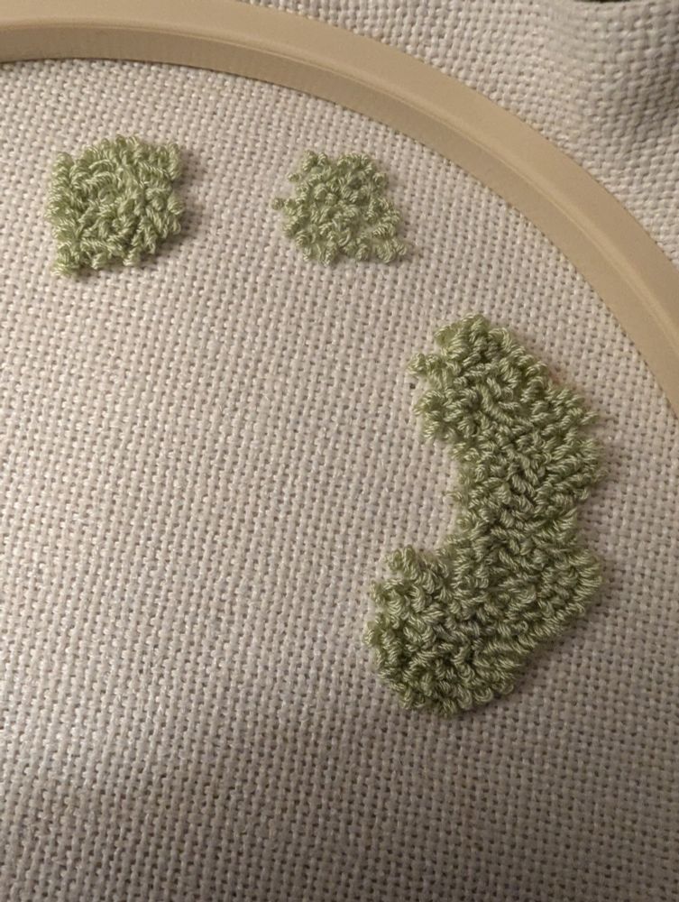 White fabric in an embroidery hoop with a few irregular patches of light green punch needle embroidery completed. No image is suggested by the shapes. 