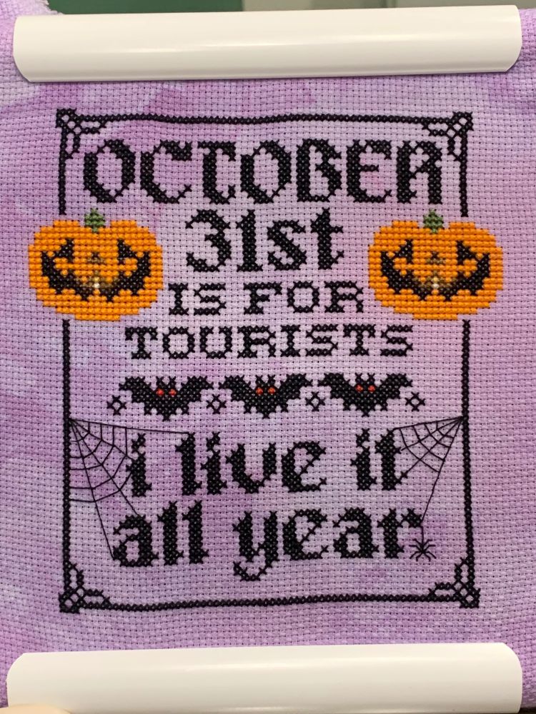 Finished cross stitch. Purple fabric, two orange jack-o-lanterns, three bats, some spider webs and black gothic text that reads “October 31st is for tourists. I live it all year.”